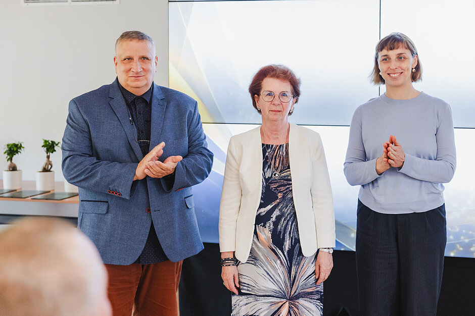 Zdjęcie przedstawia trzy osoby stojące obok siebie w jasnym pomieszczeniu, z których dwie klaszczą w dłonie. Cała grupa pozuje przed dużym ekranem, co sugeruje ich aktywny udział w oficjalnej uroczystości lub gali. 