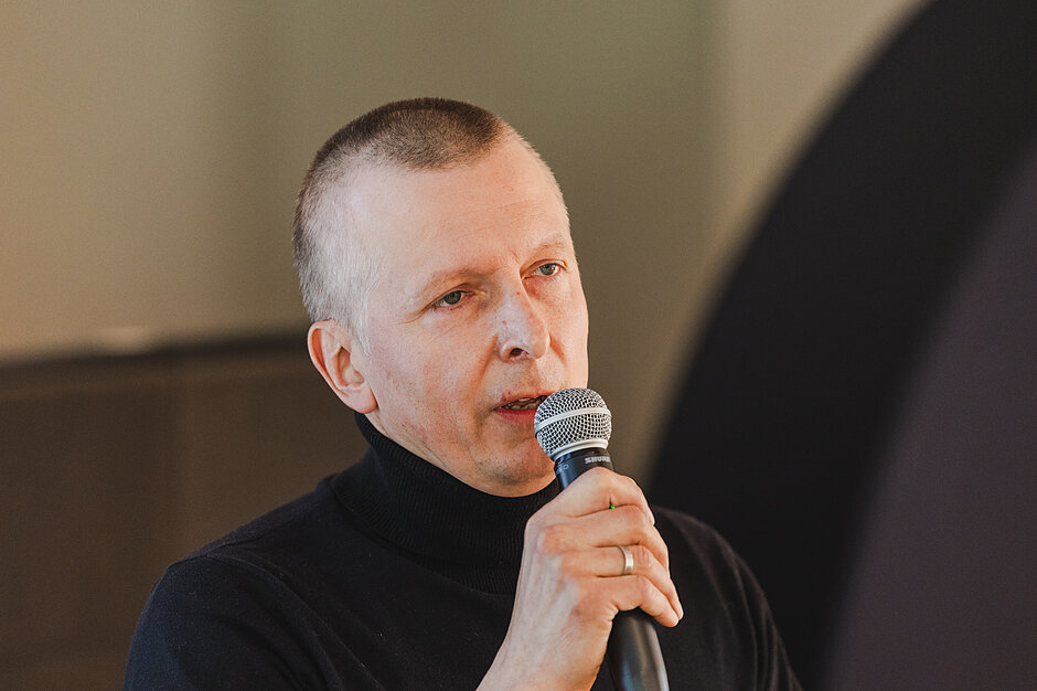 Na zbliżeniu widać mężczyznę w czarnym golfie, który z uwagą przemawia do trzymanego w dłoni mikrofonu. Jego postawa sugeruje, że występuje on w roli prelegenta lub prowadzącego podczas opisywanego wcześniej wydarzenia.