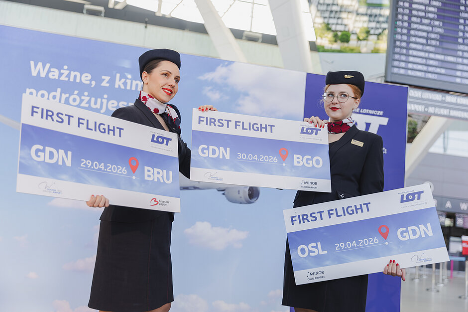 Zdjęcie przedstawia dwie uśmiechnięte stewardessy linii lotniczych LOT trzymające pamiątkowe tablice upamiętniające pierwsze loty między Gdańskiem a Brukselą, Oslo i Bergen. Wydarzenie odbywa się w terminalu lotniska, co podkreślają tablice odlotów oraz eleganckie mundury personelu pokładowego.