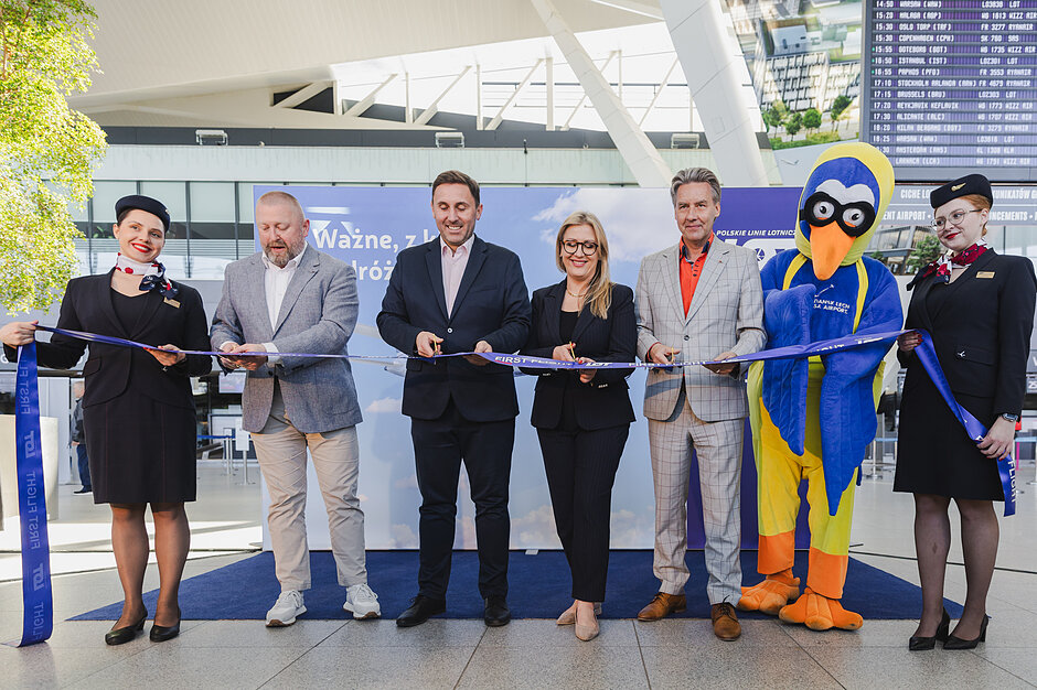 Zdjęcie dokumentuje uroczyste przecięcie wstęgi z okazji inauguracji nowych połączeń lotniczych, w którym biorą udział przedstawiciele władz i linii lotniczych LOT. Obok elegancko ubranych oficjeli i stewardess pozuje maskotka lotniska, podkreślając radosny i promocyjny charakter wydarzenia w gdańskim terminalu.