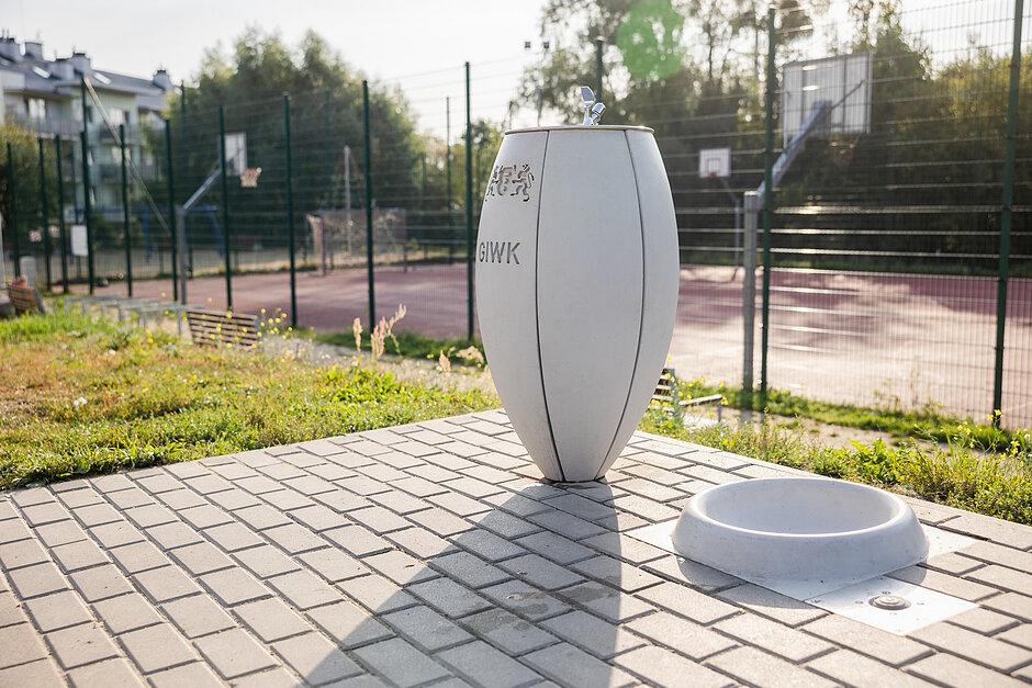 Zdjęcie przedstawia nowoczesne, białe poidełko miejskie z logo GIWK oraz znajdujący się tuż obok niski zbiornik na wodę przeznaczony dla zwierząt. Całość jest usytuowana na wybrukowanym placu tuż przy ogrodzonym boisku sportowym.