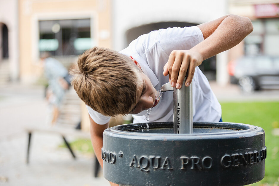 Zdjęcie przedstawia chłopca pijącego wodę bezpośrednio z nowoczesnego poidełka miejskiego z napisem „tap water”. Urządzenie z hasłem „AQUA PRO GENERATION” jest przykładem ogólnodostępnego punktu z darmową wodą pitną w przestrzeni publicznej. 