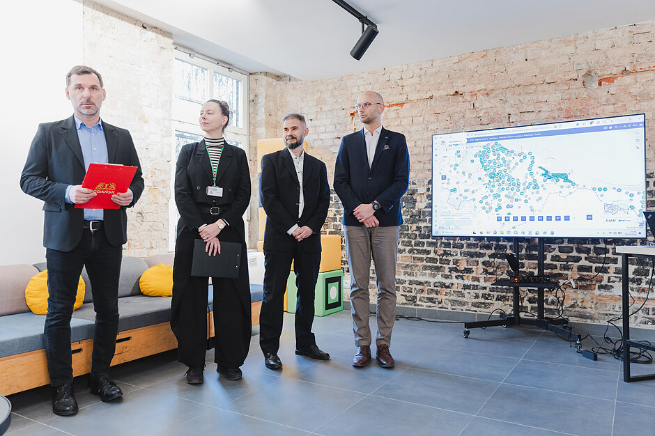 Trzech mężczyzn i jedna kobieta stoją w sali konferencyjnej, za nimi ekran z prezentacją.