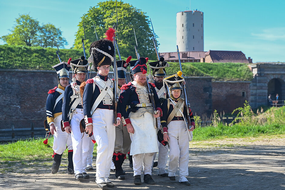 Na zdjęciu widać grupę rekonstruktorów w historycznych mundurach napoleońskich, maszerujących na tle zabytkowej Twierdzy Wisłoujście w Gdańsku. Uwagę zwracają ich bogato zdobione stroje, wysokie czapki oraz repliki dawnej broni palnej, które oddają klimat dawnych epok.