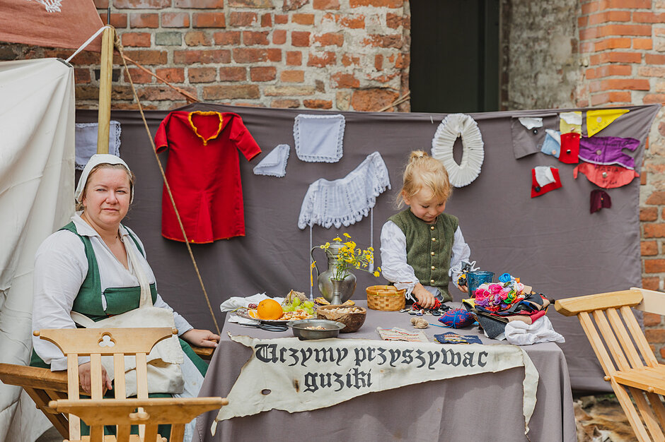 Na zdjęciu widzimy kobietę i dziecko w strojach z epoki, którzy przy stoisku z napisem „Uczymy przyszywać guziki” prezentują dawne rzemiosło. Wokół nich rozmieszczono barwne fragmenty materiałów i repliki historycznych kołnierzy, co nadaje całości charakteru edukacyjnej rekonstrukcji historycznej.