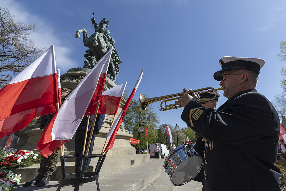 na zdjęciu z prawej żołnierz gra na trąbce, po lewej kilka białoczerwonych flag, w tle pomnik konia