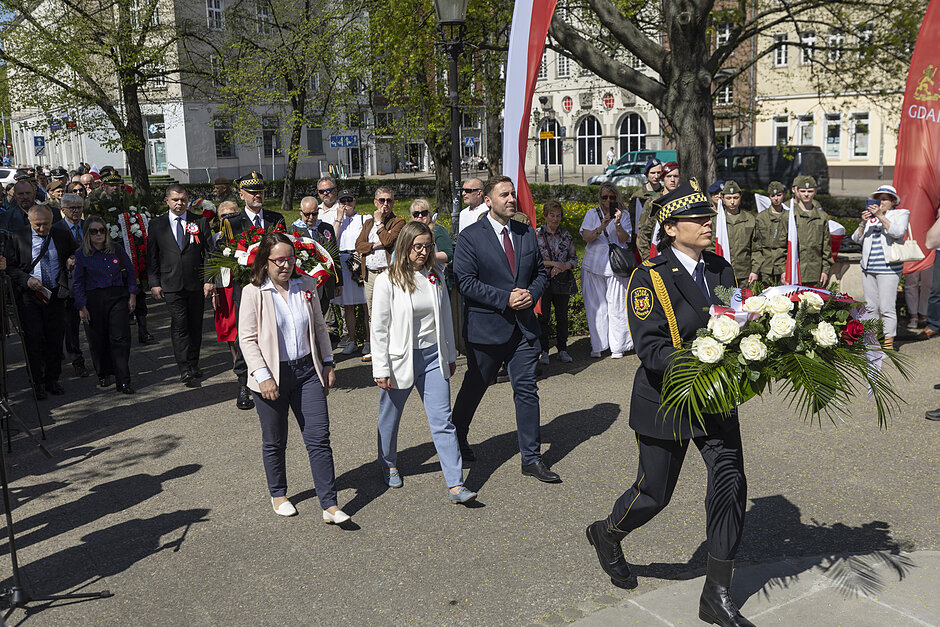 na zdjęciu trzy osoby idą w szeregu przed nimi idzie żołnierz z wieńcem