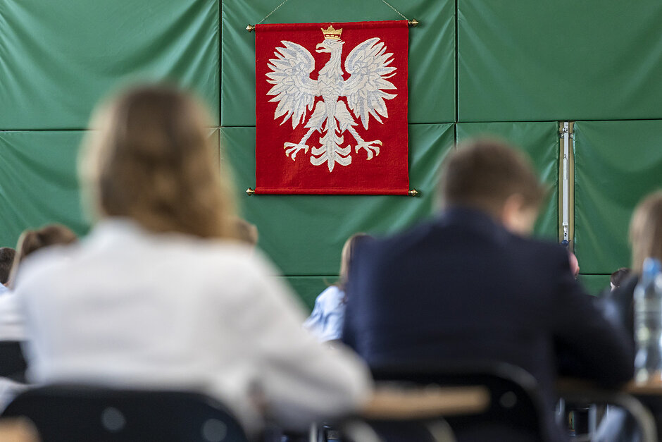 Widać od tyłu głowy osób piszących egzamin. Obiektyw aparatu wyostrzony jest na ścianę w głębi zdjęcia - na godło RP, które wisi na ścianie 