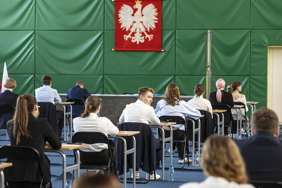 Sala gimnastyczna w szkole, przy stołach, które ustawione są w równych odstępach, siedzą uczniowie, którzy piszą egzamin