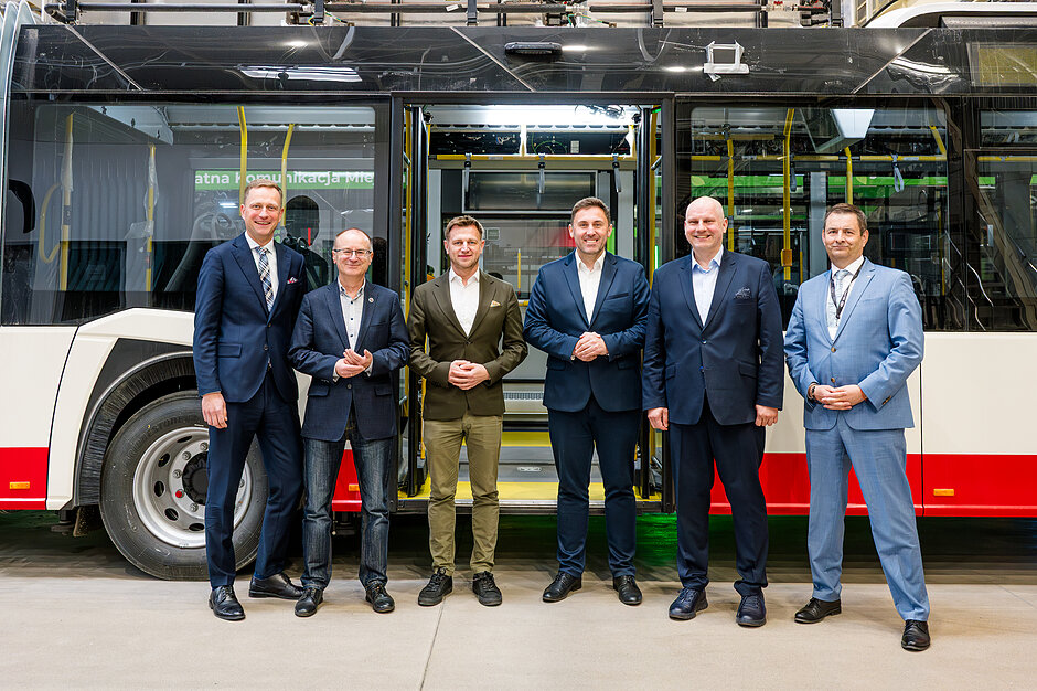 Sześciu mężczyzn pozuje wspólnie do zdjęcia, na tle autobusów w hali produkcyjnej 