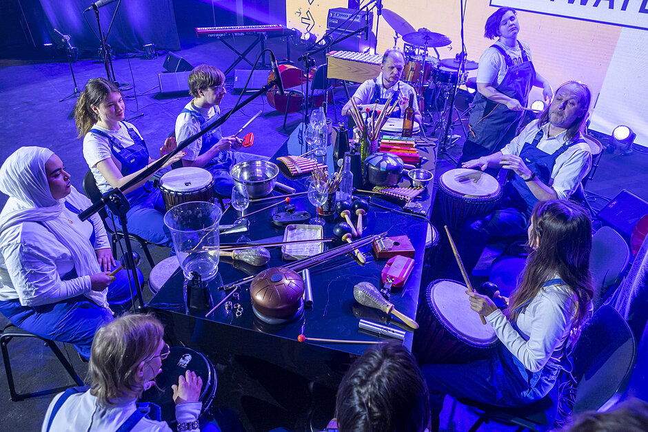 Grupa osób z instrumentami muzycznymi, siedzą przy stole na scenie