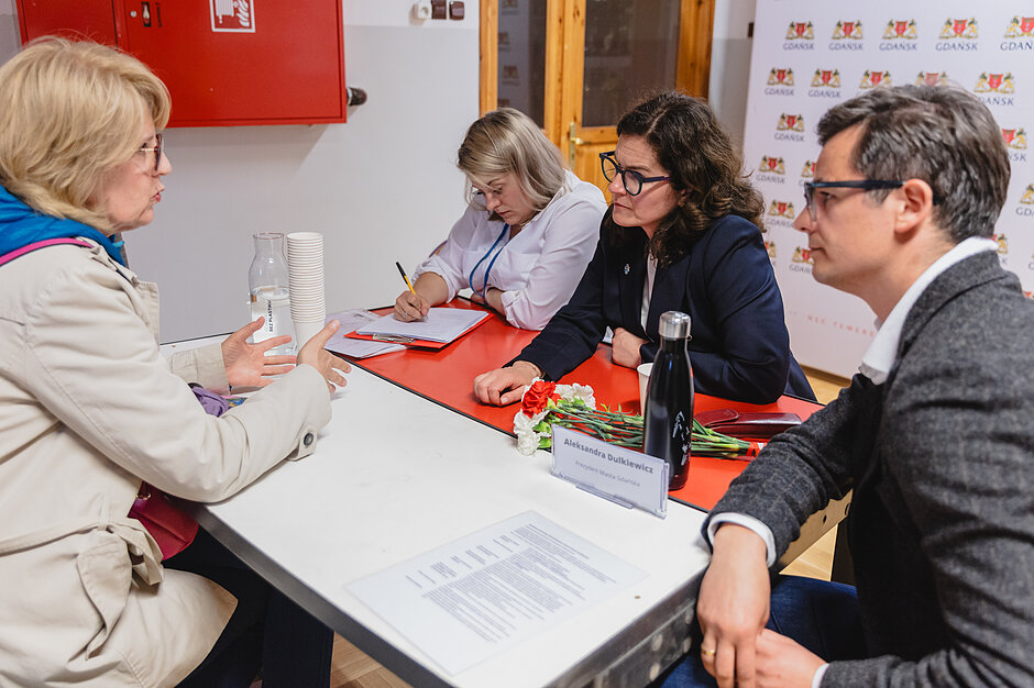 przy stole siedzą dwie kobiety przy stole, naprzeciw siebie, rozmawiają, obok siedzi trzecia kobieta która coś notuje, siedzi też mężczyzna który się temu przygląda