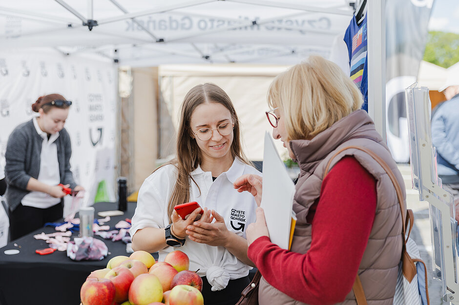 Zdjęcie przedstawia uśmiechniętą młodą kobietę w białej koszulce z logo „Czyste Miasto Gdańsk”, która pokazuje coś na telefonie starszej osobie. W tle widoczne jest stoisko promocyjne z jabłkami na pierwszym planie, co sugeruje udział w lokalnym wydarzeniu plenerowym.