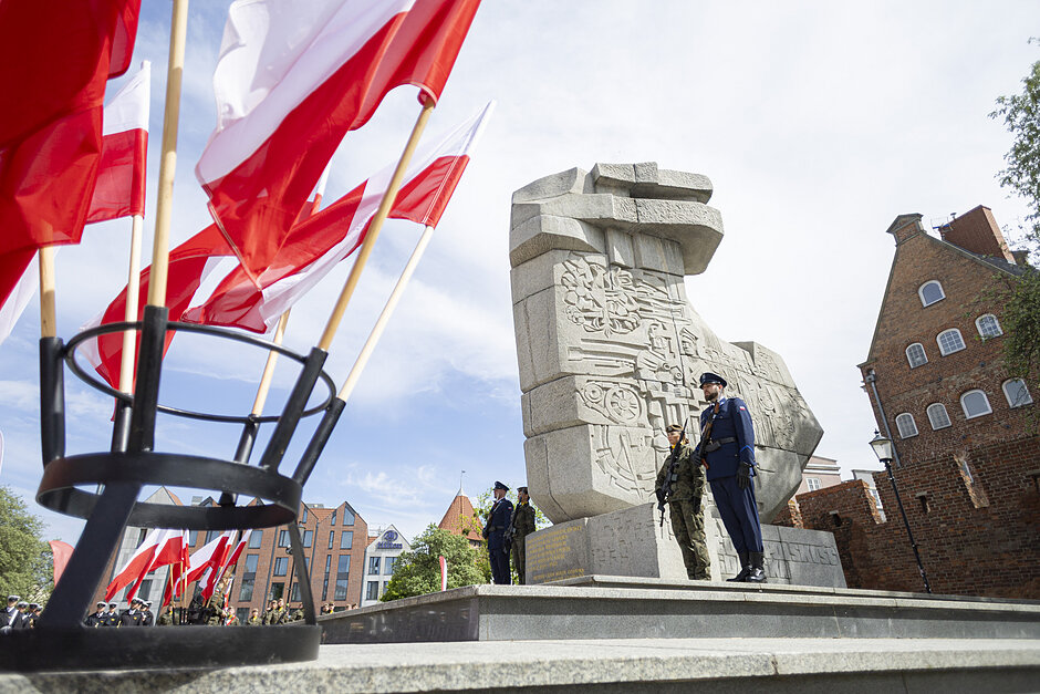 na zdjęciu w tle jasny wysoki pomnik przy którym stoi żołnierz, po lewej widać kilka stojących biało czerwonych flag