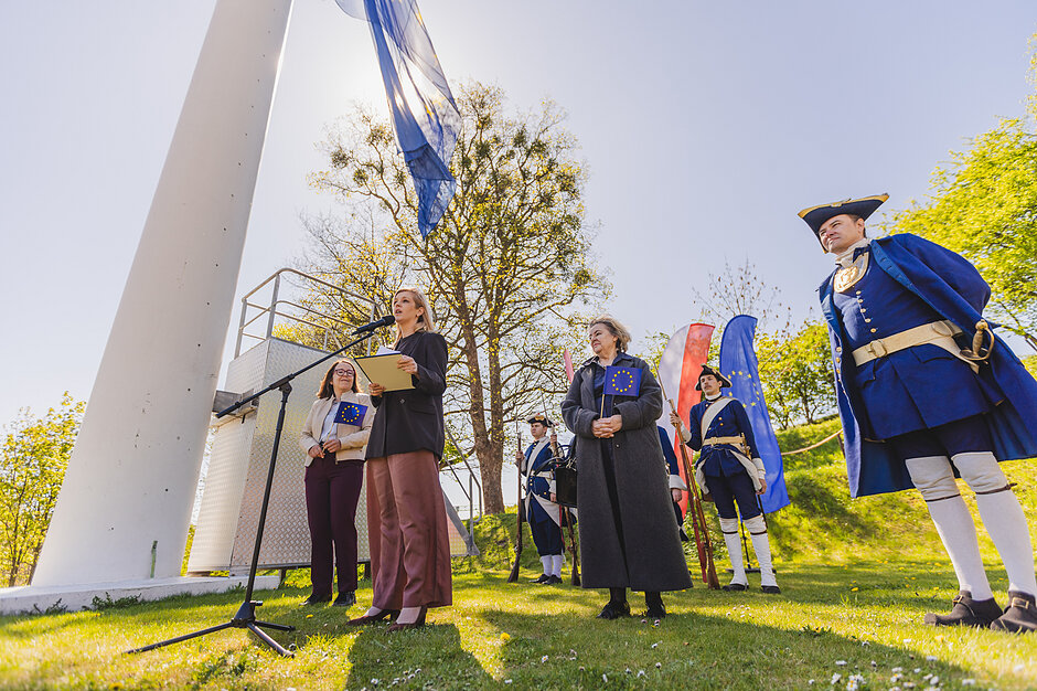 Kobieta przemawia do mikrofonu w parku miejskim pod masztem, na który podnoszona jest flaga Europy. Obok niej stoją dwie inne kobiety oraz kilka osób w mundurach z dawnej epoki.