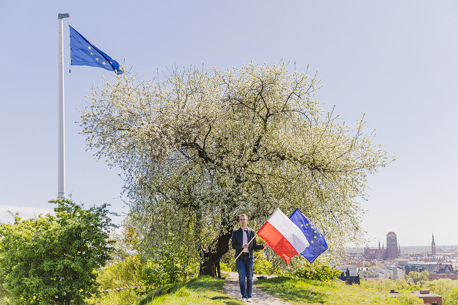 Flaga Europy powiewa na wysokim maszcie na górze nad starym miastem. Obok masztu kwitnące drzewko owocowe, pod którym idzie młody człowiek z flagami Polski i Europy.