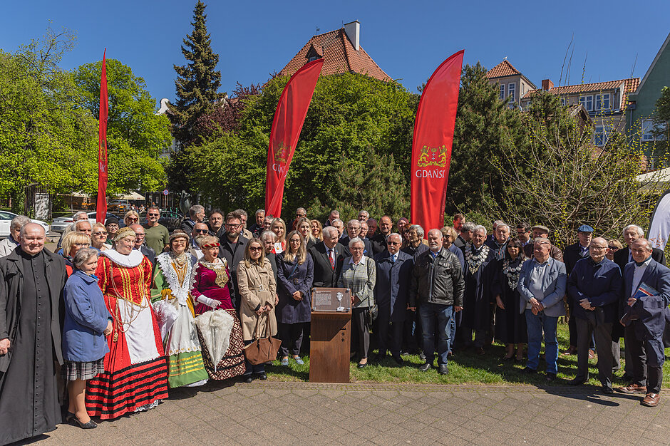 Kilkadziesiąt osób pozuje do zdjęcia w miejskim parku przy odsłoniętej tablicy pamiątkowej.