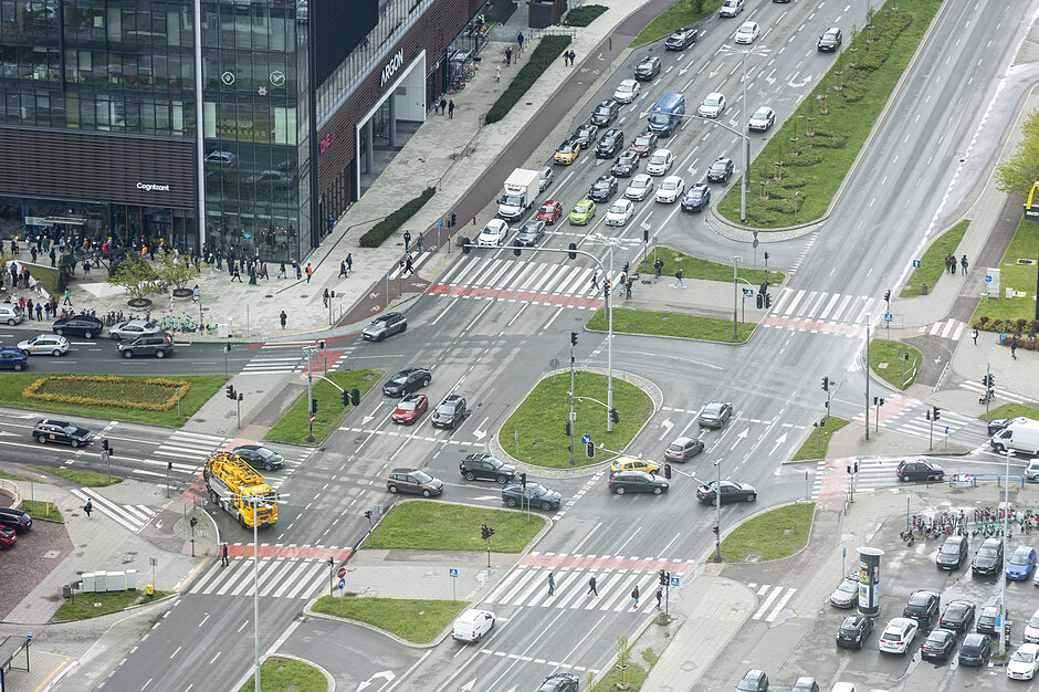 Zdjęcie przedstawia widok z góry na ruchliwe skrzyżowanie w gdańskiej Oliwie, z widocznym intensywnym ruchem samochodowym i pieszym. W tle widać nowoczesny biurowiec Argon, będący częścią kompleksu biznesowego Alchemia.