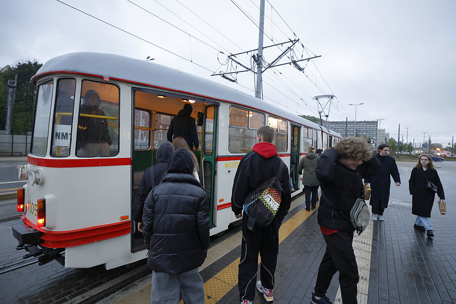 Na zdjęciu widać pasażerów wsiadających do zabytkowego, biało-czerwonego tramwaju o numerze 1105 stojącego na przystanku. Oznaczenie linii NM1 sugeruje, że jest to specjalny przejazd historyczny odbywający się w pochmurny dzień.