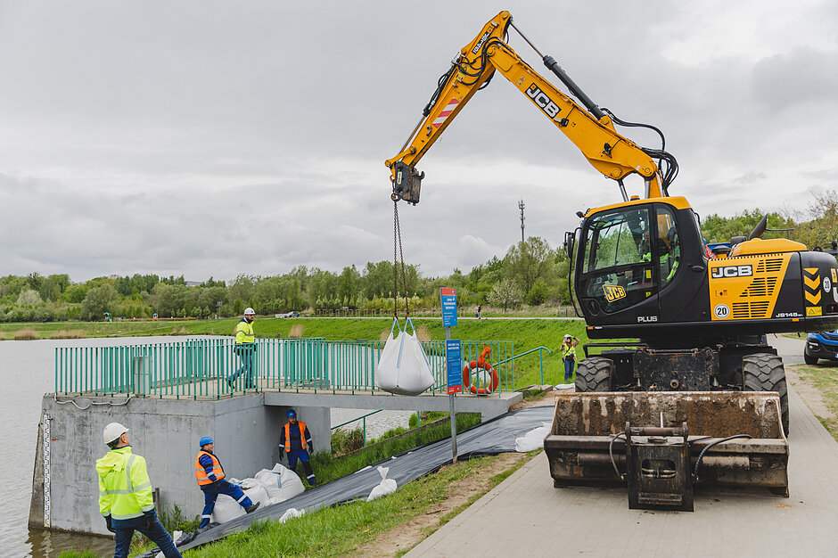 Przy zbiorniku retencyjnym pracują ludzie, dźwig transportuje wielkie worki z piaskiem.
