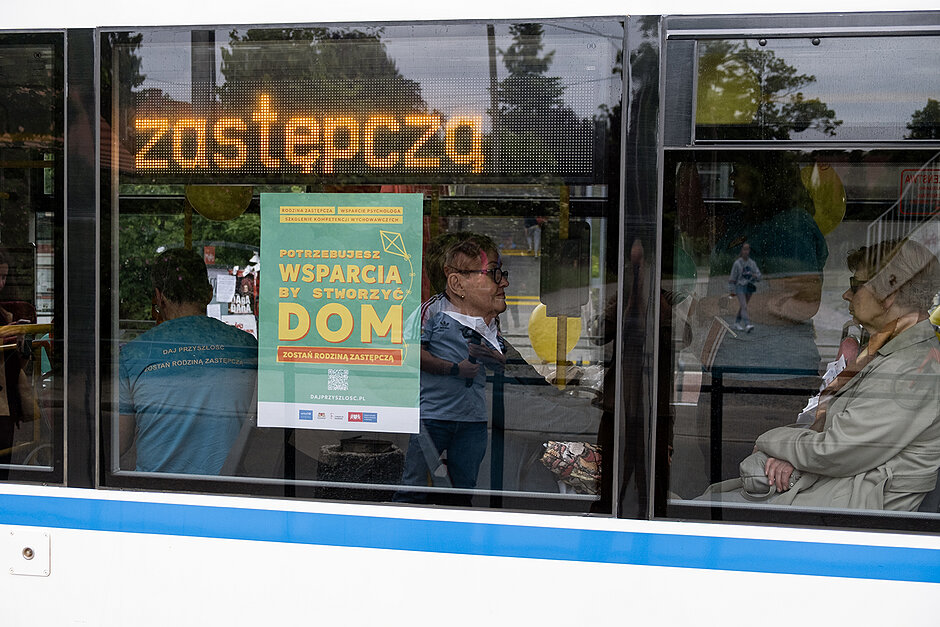 Na oknie tramwaju plakat promujący rodziny zastępcze, w środku siedzą ludzie.
