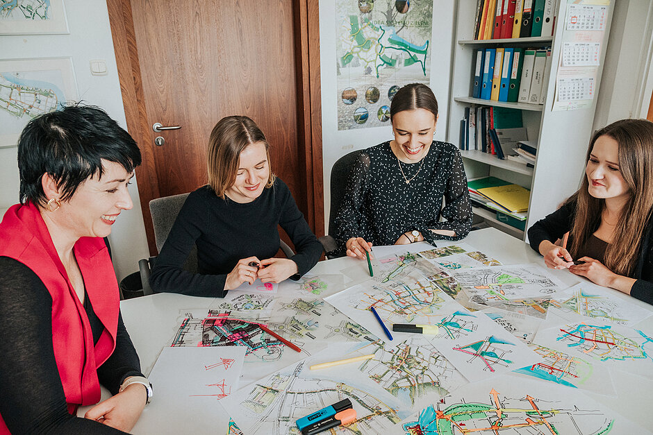 Cztery kobiety siedzą przy stole w biurze i z uśmiechem analizują kolorowe plany urbanistyczne lub architektoniczne. Na blacie leżą liczne szkice, mapy oraz mazaki, które służą im do wspólnej pracy nad projektem.