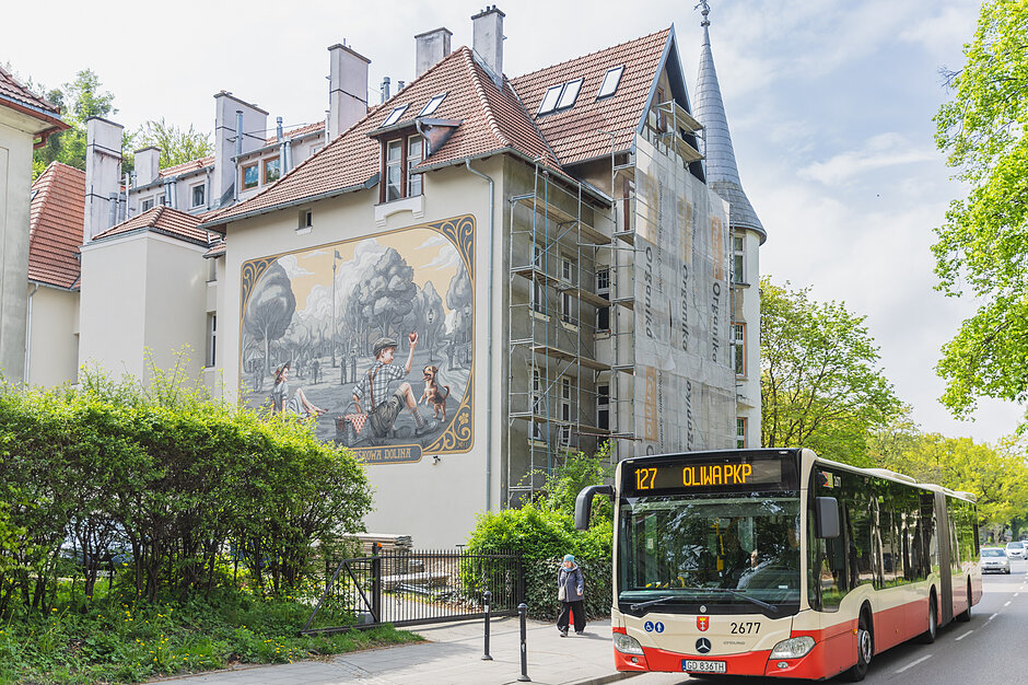 Zdjęcie przedstawia zabytkową kamienicę w Gdańsku-Wrzeszczu, na której bocznej ścianie widnieje malowniczy mural nawiązujący do dawnego życia Jaśkowej Doliny. Na pierwszym planie, obok budynku pokrytego częściowo rusztowaniem, po ulicy porusza się miejski autobus linii 127 zmierzający w kierunku Oliwy PKP.