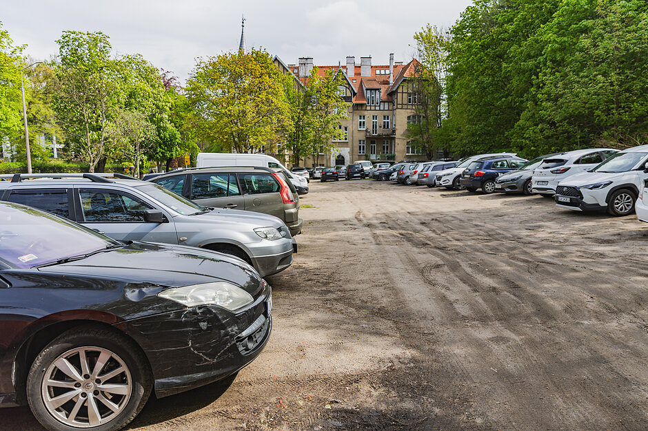 Współczesne zdjęcie przedstawia ten sam obszar dawnej Łąki Festynowej, który obecnie służy jako gruntowy, nieutwardzony parking dla samochodów. W tle, za rzędami aut i zielonymi drzewami, wciąż widoczna jest ta sama historyczna kamienica z charakterystyczną wieżyczką przy ulicy Jaśkowa Dolina.