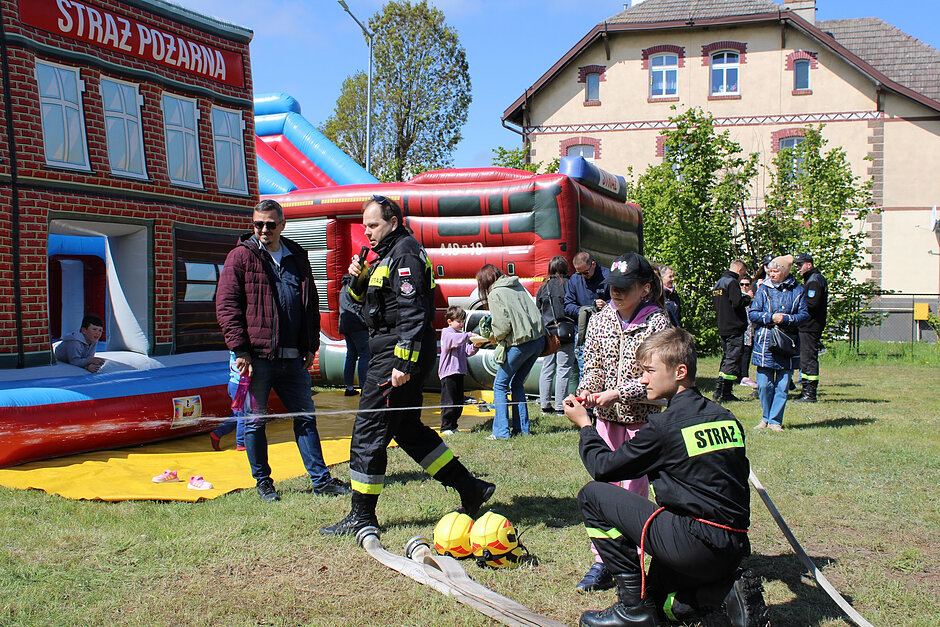 Zdjęcie przedstawia piknik lub festyn rodzinny o tematyce strażackiej, na którym dzieci bawią się w pobliżu dmuchanego zamku przypominającego budynek straży. Umundurowani strażacy pomagają uczestnikom obsługiwać wąż pożarniczy, a w tle widać zgromadzonych ludzi oraz budynek mieszkalny.