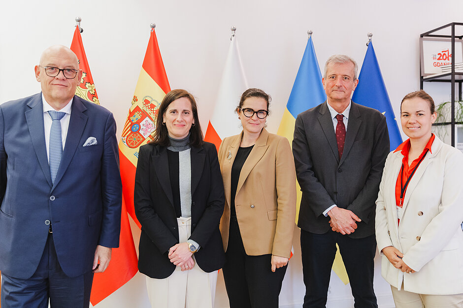 Pięć osób wspólnie pozuje do zdjęcia na tle czterech flag: polskiej, hiszpańskiej, gdańskiej i europejskiej