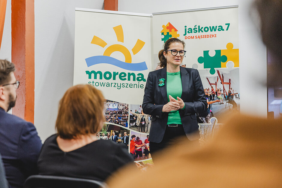 Zdjęcie przedstawia kobietę w okularach i eleganckiej marynarce, która przemawia do publiczności zgromadzonej na spotkaniu. W tle widoczne są dwa banery promujące Stowarzyszenie Morena  oraz Dom Sąsiedzki  Jaśkowa 7 .