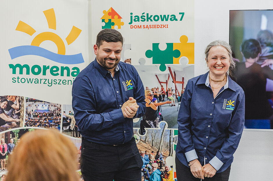 Zdjęcie przedstawia uśmiechniętą kobietę i mężczyznę w ciemnoniebieskich koszulach z logo stowarzyszenia, stojących przed publicznością. W tle znajdują się banery informacyjne Domu Sąsiedzkiego „Jaśkowa 7” oraz „Stowarzyszenia Morena”.