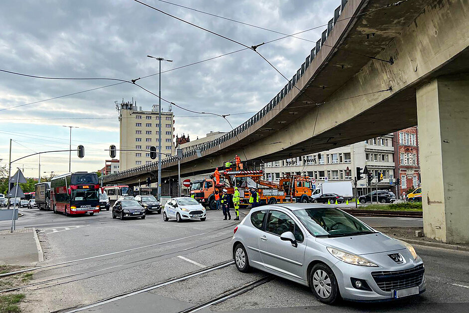 zdjęcie ulicy, po której jadą auta i autobusy, na środku widoczna ekipa naprawiająca trakcję tramwajową