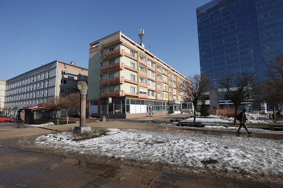 Zdjęcie przedstawia miejski krajobraz w zimowej lub wczesnowiosennej scenerii, gdzie topniejący śnieg tworzy kałuże na chodniku. W tle widoczne są zróżnicowane architektonicznie budynki, w tym wielopiętrowy blok mieszkalny z balkonami oraz nowoczesny, szklany wieżowiec.