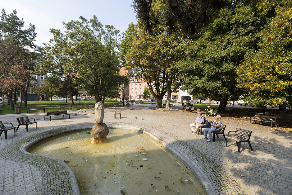 Zdjęcie przedstawia niewielki miejski skwer z fontanną o nieregularnym kształcie, w której centrum umieszczono kamienną rzeźbę. Wokół wody rozstawione są ławki, na których w otoczeniu bujnej zieleni drzew odpoczywają przechodnie.