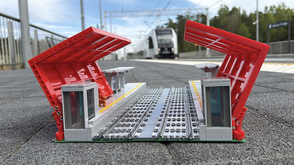 Zdjęcie przedstawia precyzyjnie wykonany model stacji kolejowej z klocków, odwzorowujący charakterystyczne czerwone wiaty Pomorskiej Kolei Metropolitalnej. Miniatura została ustawiona na prawdziwym peronie przystanku Gdańsk Firoga, a w tle widać nadjeżdżający, pełnowymiarowy pociąg.