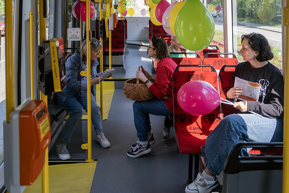 Wnętrze tramwaju z balonami, w którym rozmawiają ludzie.
