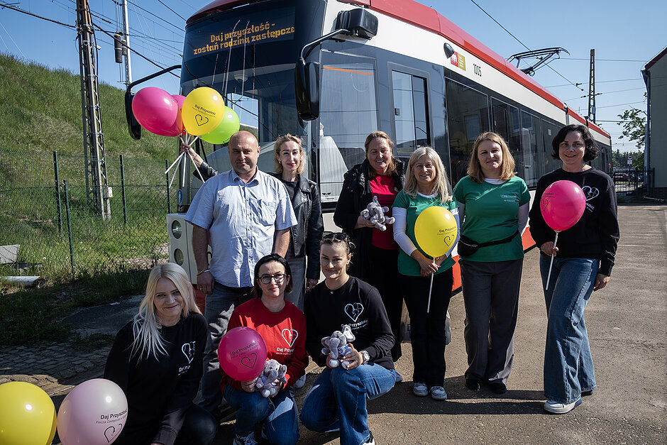 Kilka osób pozuje do zdjęcia z balonami przed tramwajem.