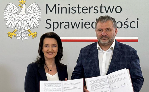 Kobieta i mężczyzna stoją obok siebie w Ministerstwie Sprawiedliwości i trzymają otwarty, podpisany dokument. W tle widoczne jest godło Polski oraz oficjalne logo ministerstwa.
