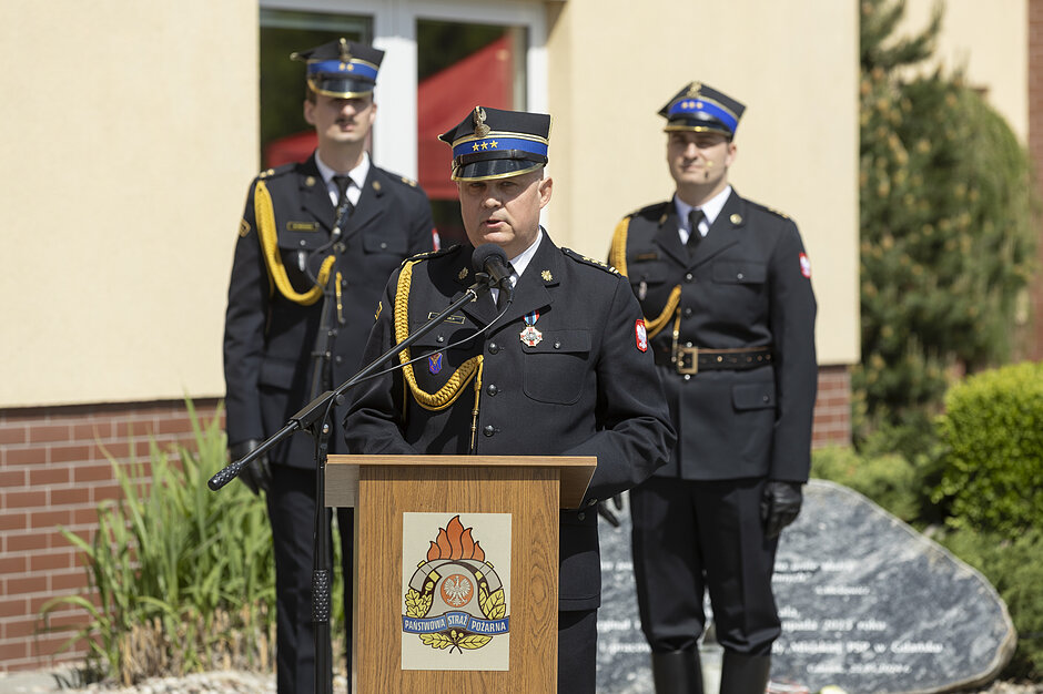 Fotografia przedstawia oficera Państwowej Straży Pożarnej w mundurze galowym, który przemawia do zgromadzonych stojąc przy drewnianej mównicy z emblematem formacji. W tle, przed budynkiem, asystuje mu dwóch innych umundurowanych strażaków w czapkach rogatywkach.