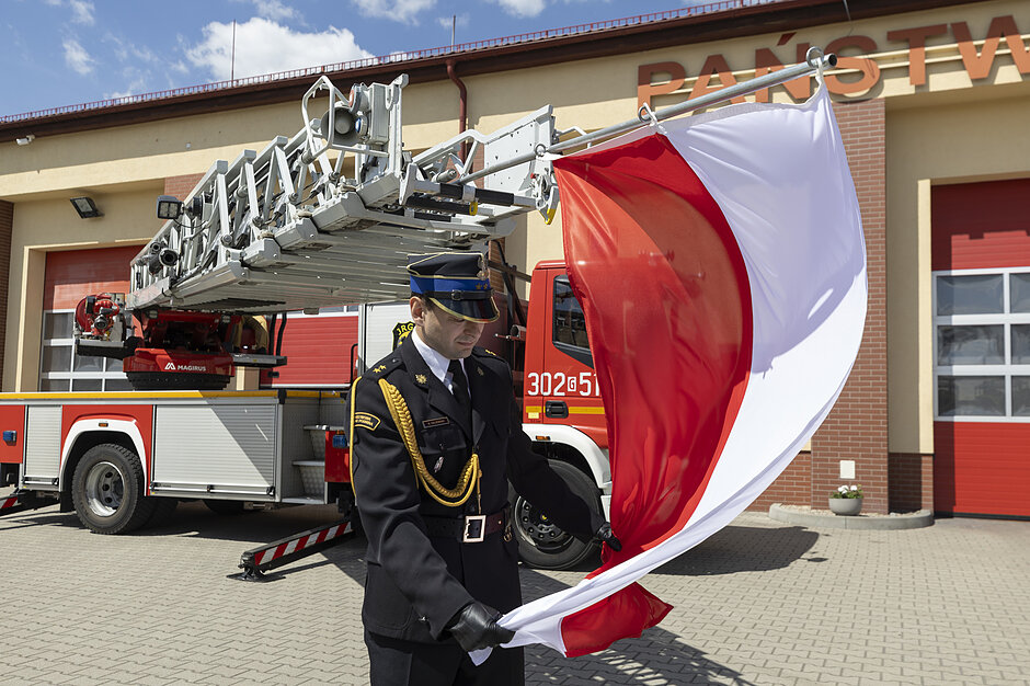 Zdjęcie przedstawia strażaka w mundurze galowym, który rozwija lub składa dużą biało-czerwoną flagę Polski. W tle widoczny jest plac przed budynkiem jednostki oraz wóz strażacki wyposażony w drabinę mechaniczną.