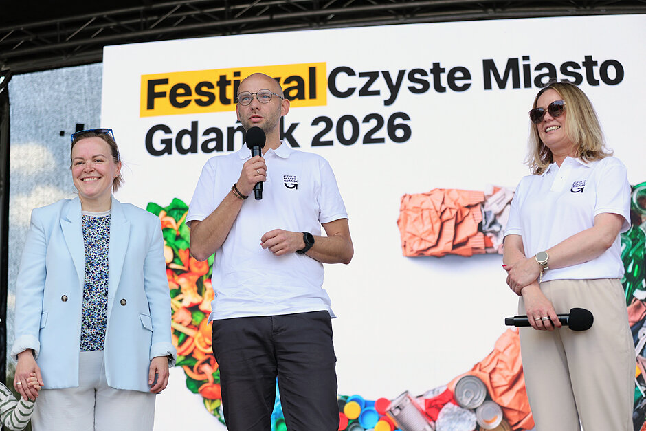 Trzy osoby na scenie, dwie stoją obok siebie po lewej, trzecia - po prawej, z mikrofonem