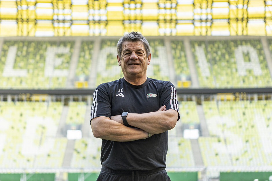 Mężczyzna lat około 60, w dresie, pozuje do zdjęcia na tle trybun stadionu