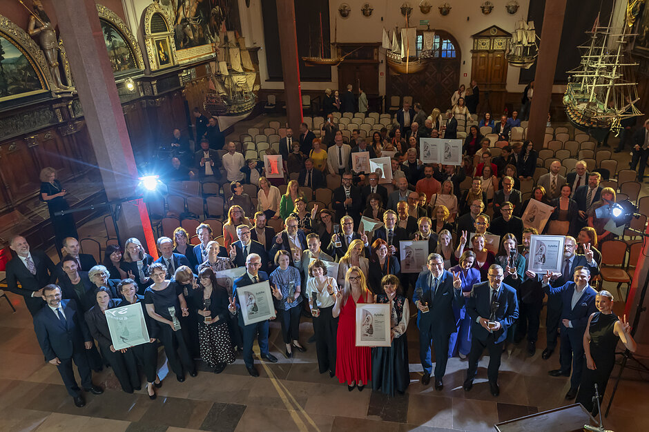 Zdjęcie przedstawia dużą grupę elegancko ubranych osób zgromadzonych w zabytkowym, bogato zdobionym wnętrzu, przypominającym muzeum lub salę ceremonialną z modelami statków pod sufitem. Wielu uczestników spotkania z uśmiechem prezentuje otrzymane dyplomy oraz statuetki, pozując wspólnie do pamiątkowej fotografii zrobionej z podwyższenia.