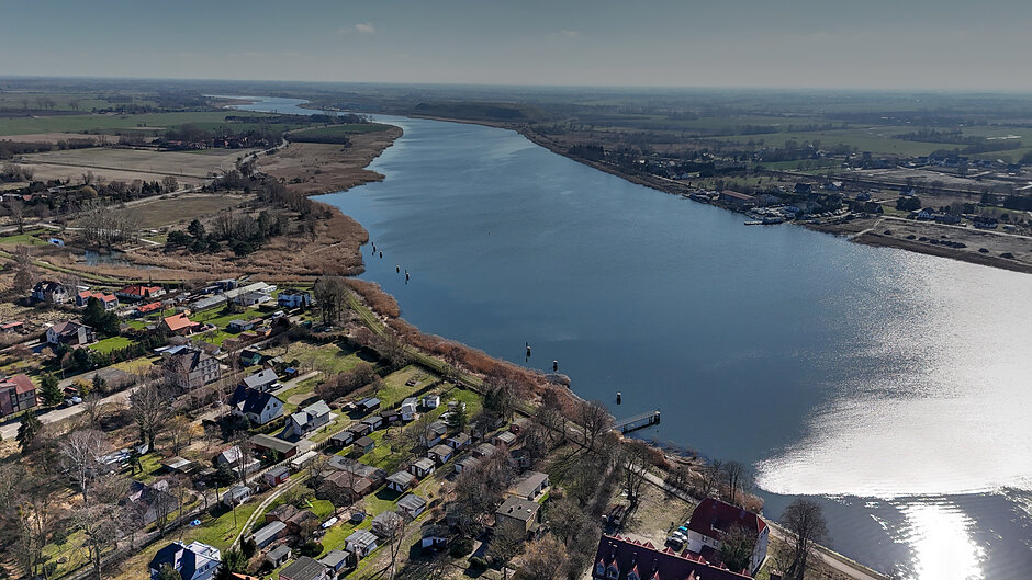 Zdjęcie przedstawia panoramiczny widok z góry na szerokie, spokojne jezioro lub rzekę otoczoną naturalną roślinnością i trzcinowiskami. Na pierwszym planie i wzdłuż brzegu widoczna jest niska zabudowa rekreacyjna, domki letniskowe oraz rozległe pola uprawne w oddali.