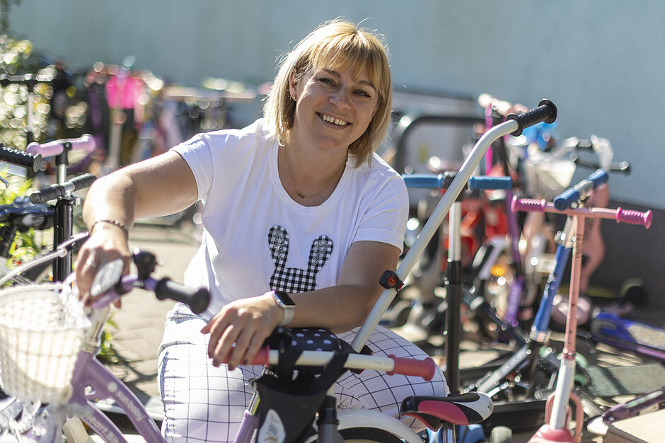 Uśmiechnięta kobieta w białej koszulce kuca na parkingu pośród licznych dziecięcych rowerów i hulajnóg. Trzyma dłonie na kierownicy jednego z jednośladów, a w tle widać rozmyte sylwetki kolejnych zaparkowanych pojazdów.