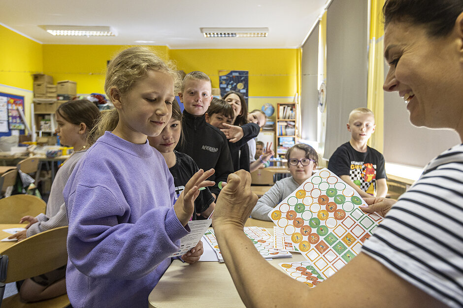 Uśmiechnięta nauczycielka rozdaje dzieciom w klasie kolorowe naklejki z rowerami, które służą do zaznaczania postępów w kampanii. Dziewczynka w fioletowej bluzie z zaciekawieniem ogląda otrzymany element, podczas gdy inne dzieci czekają w kolejce.
