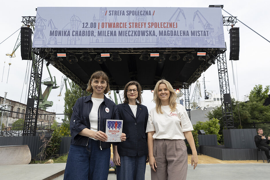 na zdjęciu pozują trzy kobiety w średnim wieku, uśmeichają się, za nimi widać dźwig stoczniowy
