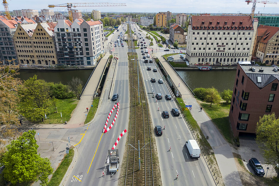 - ruchliwa ulica miejska z wieloma pasami ruchu. Ulica jest wyłożona wysokimi budynkami po obu stronach i widocznych jest kilka żurawi w tle. Po lewej stronie znajduje się rzeka z mostem przechodzącym przez nią. W centrum tej ulicy biegnie równolegle do ulicy tor kolejowy. Na torach znajdują się czerwono-białe paskowe bariery wskazujące, że droga jest zamknięta z powodu prac budowlanych lub konserwacyjnych.