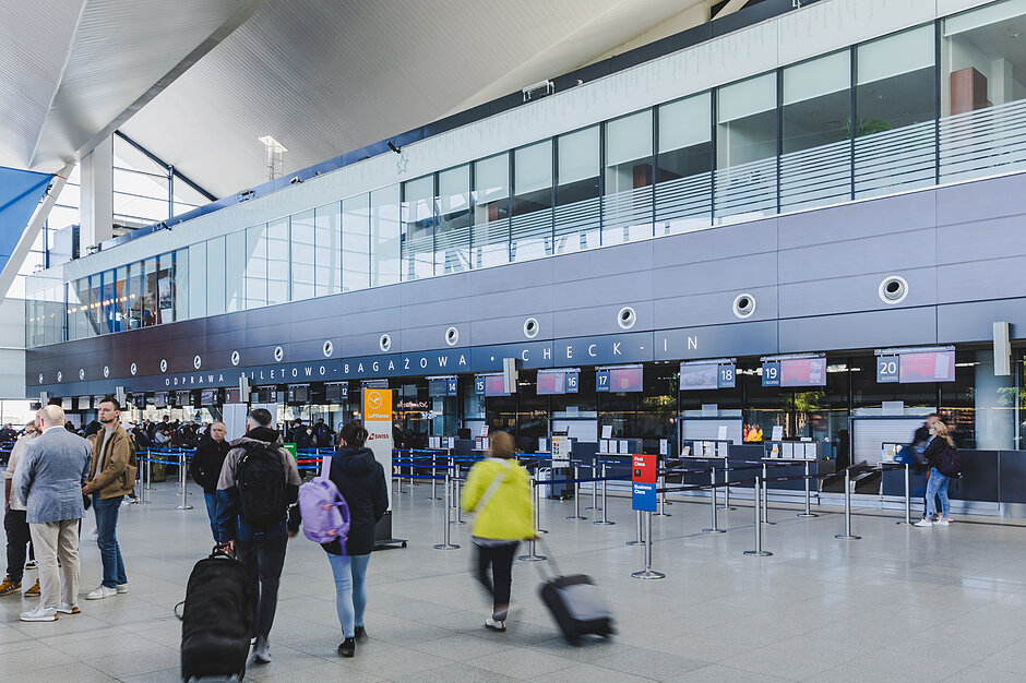 Zdjęcie przedstawia nowoczesny i przestronny terminal pasażerski lotniska w strefie odprawy biletowo-bagażowej. Widoczni są podróżni przemieszczający się z walizkami oraz stanowiska Check-in  zlokalizowane pod ciemną konstrukcją z liniami okien powyżej.